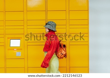 Hipster fashion young woman in bright clothes going past modern postal automatic mail terminal with self service device for receiving an order. Electronic locker for storing parcels. Selective focus. Royalty-Free Stock Photo #2212146523