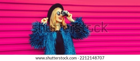 Portrait of stylish happy smiling young woman photographer with film camera taking picture wearing blue fur coat, black round hat on pink background