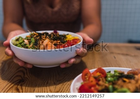 Hands of unrecognizable woman holding and displaying a poke bowl on a wooden table. Colorful picture of typical hawaiian food.