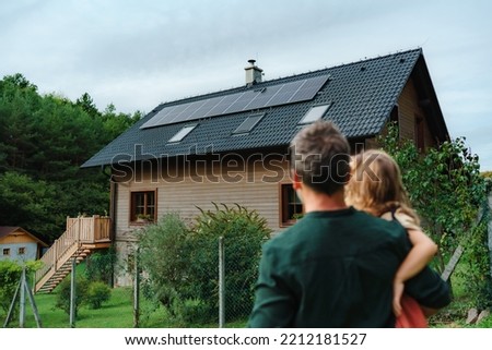 Rear view of dad holding her little girl in arms and looking at their house with installed solar panels. Alternative energy, saving resources and sustainable lifestyle concept. Royalty-Free Stock Photo #2212181527