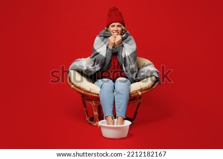 Full body frozen sad young woman wear grey plaid scarf hat sit in chair soaring feet isolated on plain red background studio portrait. Healthy lifestyle ill sick disease treatment cold season concept Royalty-Free Stock Photo #2212182167
