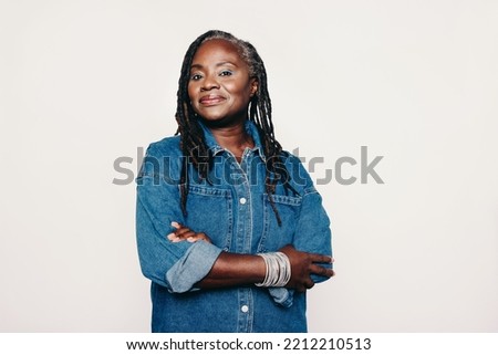 Stylish mature woman looking at the camera while standing against a grey background with her arms crossed. Fashionable woman with dreadlocks wearing a denim jacket and make-up. Royalty-Free Stock Photo #2212210513