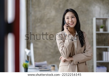 Successful Asian businesswoman using smartphone to chat with clients busy working at desk in modern office. looking at the computer screen with a smile