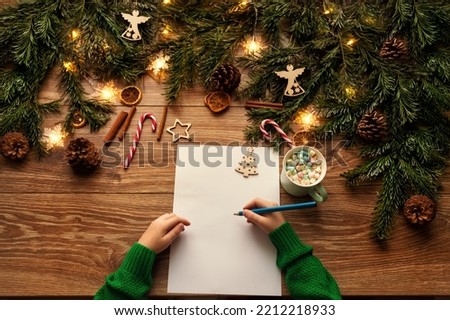 Letter to Santa. Christmas Child Hands Close up writing Xmas Gifts Shopping Wish List over brown Wooden Table. Wood Background with Fir Tree Branch decorated Christmas Lights and Toys Royalty-Free Stock Photo #2212218933