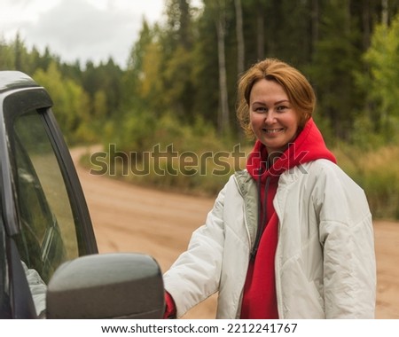 a woman with red hair in a white jacket opening the car door against the background of a forest road and forest. the concept of travel and tourism.