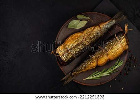 two hot smoked herrings with ropes and spices lie side by side on a clay plate on a black board and a black background. view from above. dark still life with copy space