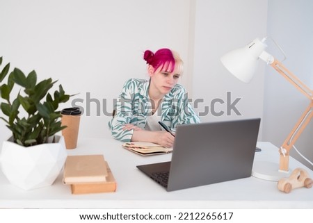 Young pink haired blonde woman sitting at table in office and looking at laptop screen and writing in notebook