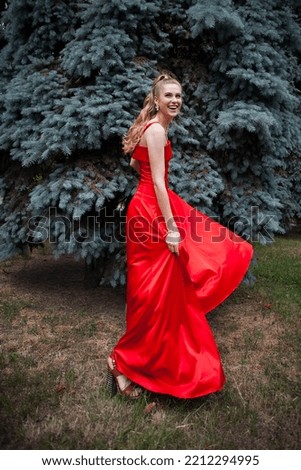 A cute smiling young girl with blond hair and beautiful makeup, in a long red dress, stands against the backdrop of trees in the park.