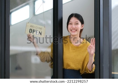 Young Asia manager girl changing a sign from closed to open sign on door cafe looking outside waiting for clients after lockdown. Owner small business, food and drink, business reopen again concept