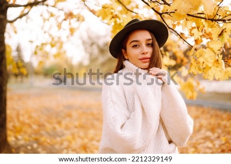 Portrait of attractive stylish smiling woman walking in park dressed in warm white sweater autumn trendy fashion, street style wearing hat.