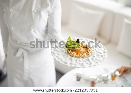 Chef holds a plate with delicious vegetarian meal decorated with flower made of avocado at restaurant of haught cuisine, close-up on dish Royalty-Free Stock Photo #2212339931