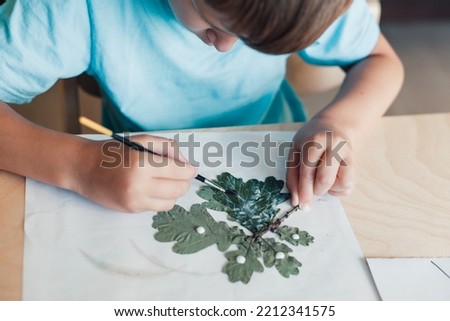 8 years old boy sitting by desk and making herbarium from oak leaves on album sheet. Autumn activities for children