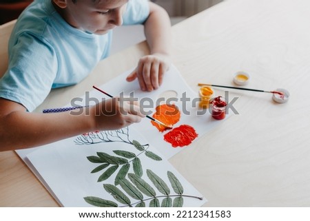 Cute child boy siting at desk and drawing rowanberries on album sheet with dry rowan leaves. Autumn activities