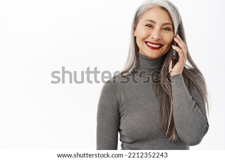 Portrait of beautiful and happy asian senior woman, middle aged lady talking on mobile phone, calling someone and smiling, standing over white background