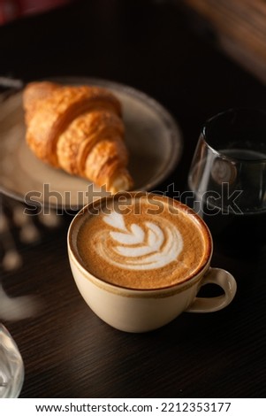 Cup of Cappuccino with beautiful cream latte art and a croissant on the side on a coffee shop table. Side view. Black Background. Royalty-Free Stock Photo #2212353177