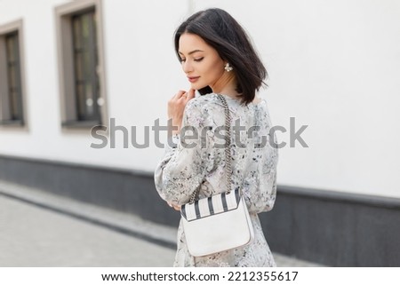 Fashionable young elegant pretty woman model with shorts hairstyle in stylish spring vintage dress with flowers print with fashion white bag walks on the city near a white wall Royalty-Free Stock Photo #2212355617