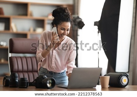 Beautiful creative young woman photographer working in office, having phone conversation with client, looking at laptop screen, set the shooting, copy space. Creative artist female business