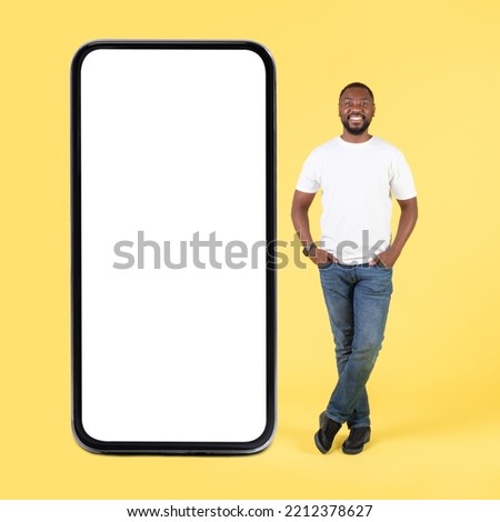 Cheerful African American Guy Standing Near Big Smartphone With Blank Screen Posing Holding Hands In Pockets, Advertising New Mobile Application Over Yellow Background. Studio Shot, Mockup