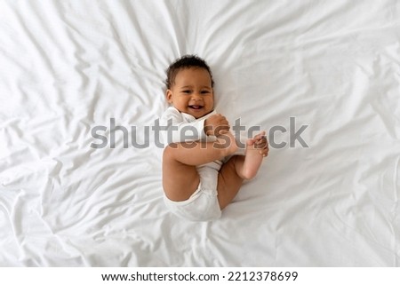 Cute Funny Little Black Baby Having Fun While Lying On Bed At Home, Adorable African American Infant Boy Playing With His Toes And Laughing While Resting In Bedroom, Top View, Copy Space Royalty-Free Stock Photo #2212378699