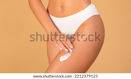 Slim woman in white panties applying moisturising body cream on her legs, using anti-cellulite lotion or sunscreen while standing on beige studio background, cropped