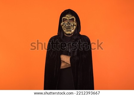 Person with skull mask and black hooded cape, with crossed arms, celebrating Halloween, on orange background. Celebration concept, All Souls' Day and All Saints' Day.
