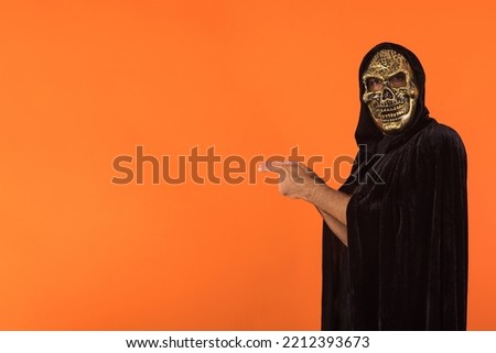 Person with skull mask and black hooded cape, pointing fingers to the side, celebrating Halloween, on orange background. Celebration concept, All Souls' Day and All Saints' Day.