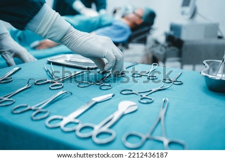 Close up surgeon doctor's hand with hygiene glove taking sterile surgical instrument tool, equipment in operating room patient in background for surgery. Hospital, medical healthcare emergency concept Royalty-Free Stock Photo #2212436187
