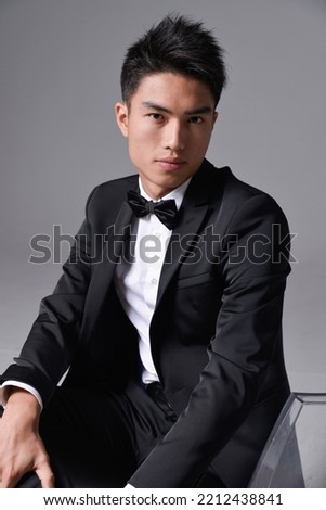 Handsome fashion model.
elegant man wear formal black suit with bow tie sitting chair on gray background 
