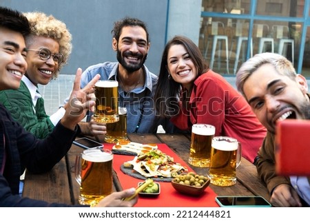 Multiracial friends take selfie with mobile phone while enjoying cool beer and food in a bar outdoors.