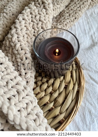 A black lighted candle in glass on a rattan stand and a plush beige plaid on the bed in the room. Selective focus. Create a cozy atmosphere by adding candles to your home decor. Flatlay. Royalty-Free Stock Photo #2212449403