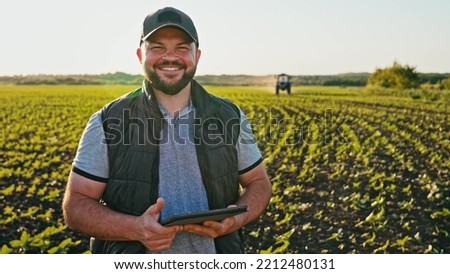 Happy farmer stands and smile holds tablet in his hands against background of working tractor in field. Concept ecology, transport, outdoor nature, clean air, food. Natural production bio product. Royalty-Free Stock Photo #2212480131
