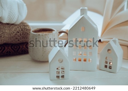A small candlestick of a Christmas house with a burning candle inside on the background of book and winter sweaters on the window.Concept Real estate,holiday.Christmas composition.Winter cozy interior