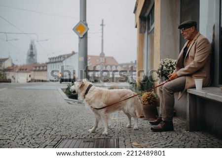 Unhappy senior man sitting on bench in front of store and waiting somebody wih his big white dog.