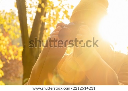 A woman photographer in a brown coat takes a photo on a retro camera in an autumn forest against sunlight, side view. 