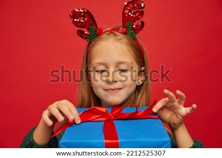Smiling funny child (kid, girl) in Reindeer Hairband opening Christmas gifts. New year  concept. Shooting on red background