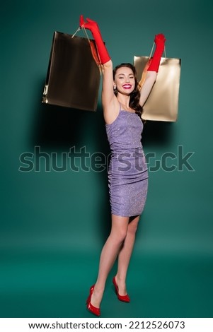 full length of excited woman in red gloves and purple dress holding shopping bags on green