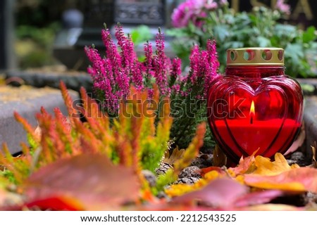 A red heart-shaped candle on a grave  in a cemetery on an autumn day. All Saints Day. Copy space, shallow depth of field. Royalty-Free Stock Photo #2212543525