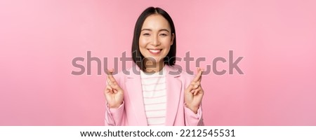 Happy asian businesswoman cross fingers for good luck, wishing, praying and hoping, smiling at camera, standing in suit over pink background