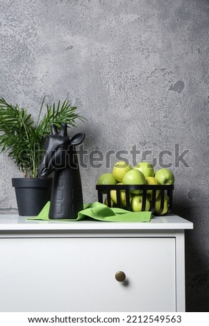 Stylish decor, basket with apples and houseplant on chest of drawers near grey wall indoors, space for text. Interior design