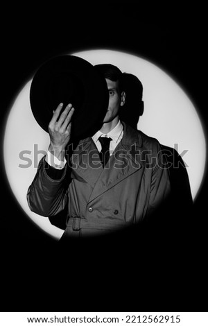 A dark silhouette of a male detective in a coat and hat in the noir style. A dramatic portrait in the style of detective films of the 1950s. The silhouette of a spy in a circle of light, like Agent Royalty-Free Stock Photo #2212562915