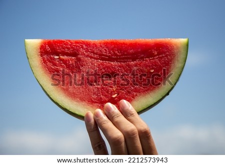 Fresh watermelon slice with sky in the background held in left hand Royalty-Free Stock Photo #2212573943