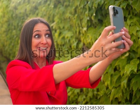 Positive adult woman with makeup in red dress standing near wall with green leaves and taking selfie while showing tongue in garden