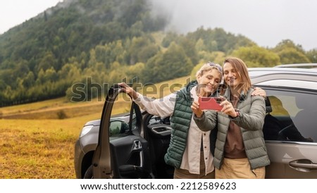 Two happy adult women mother and daughter traveling together by car taking selfie standing next to the car