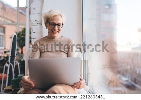 Smiling middle aged female freelancer wearing eyeglasses sitting on windowsill and looking at screen of laptop while working on remote project