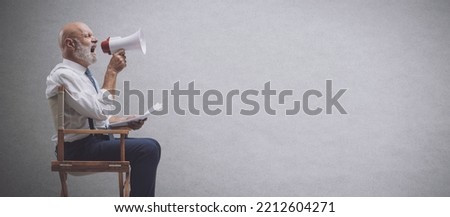 Confident professional film director sitting on the director's chair and shouting with a megaphone, film industry concept Royalty-Free Stock Photo #2212604271