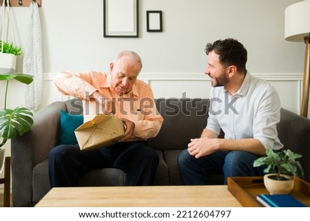 Senior caucasian man sitting on the sofa at home looking happy while opening a birthday present from his son