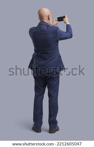 Corporate businessman taking pictures and sharing online with his smartphone, back view, full-length portrait