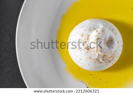 Italian mozzarella, sprinkled with herbs, lies on a white, ceramic plate, on which olive oil is spilled. Nearby is a leaf of lettuce. The plate stands on a gray, stone background.