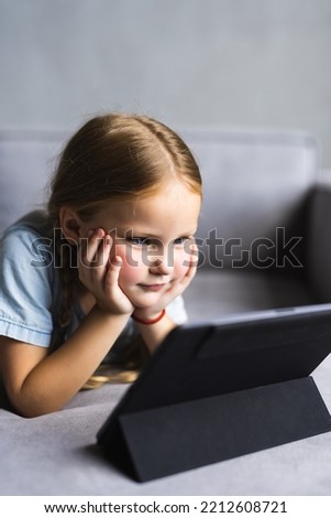 Focused cute preschooler girl watching movie, animated films, cartoons on tablet. Child girl lying on couch in living room, using digital pad for video call, holding device close to face