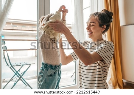 Loving mother dressing her daughter by the window Royalty-Free Stock Photo #2212615449